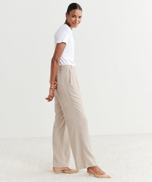 A person wearing a white t-shirt, Jones Trouser in beige, and beige flats stands sideways, smiling over their shoulder in a minimal, light-toned room.