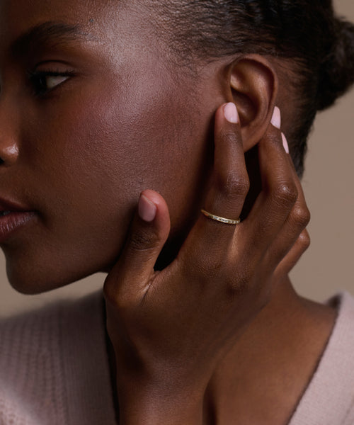 A woman in profile touches her ear, showing smooth dark skin, natural makeup, pale pink nails, and a June Baguette Band on her finger. She wears a light knit sweater.