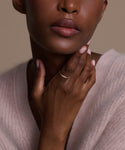 A close-up of a woman with medium-dark skin in a soft pink sweater, touching her neck. She wears light pink nail polish and the June Baguette Band on her finger. Her lips are slightly parted.