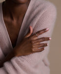 A person with medium to dark skin tone, wearing a pale pink sweater, crosses their arms to show off perfectly manicured nails and the shimmering June Baguette Band on their finger.