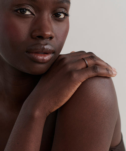 A close-up of a woman with dark skin resting her hand on her bare shoulder, highlighting the June Baguette Band—a thin 14k gold ring—on her finger as she gazes softly at the camera against a plain background.