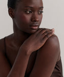 A woman with dark skin and short hair gazes to the side, gently resting her hand on her shoulder. She wears a brown camisole, a small hoop earring, and the delicate June Baguette Band in 14k gold, her expression calm and contemplative.