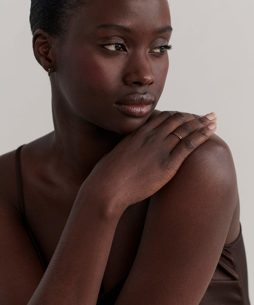 A woman with dark skin and short hair gazes to the side, gently resting her hand on her shoulder. She wears a brown camisole, a small hoop earring, and the delicate June Baguette Band in 14k gold, her expression calm and contemplative.