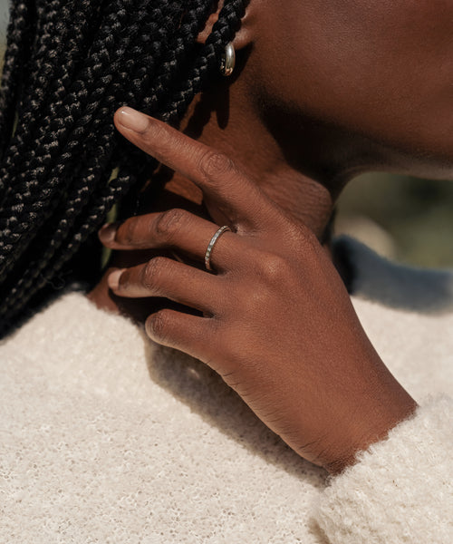 A close-up of a person with braided hair touching their face, showcasing the June Baguette Band and hoop earring, while wearing a cream textured sweater.