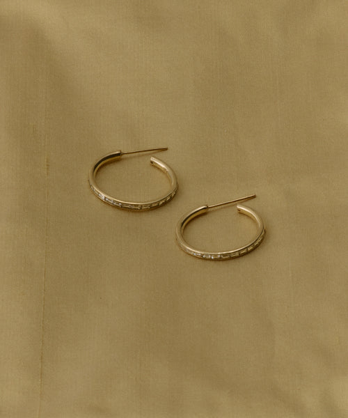 The June Baguette Hoops are small 14k gold earrings featuring engraved details, inspired by Jenni Kayne jewelry, and shown on a light beige fabric background.
