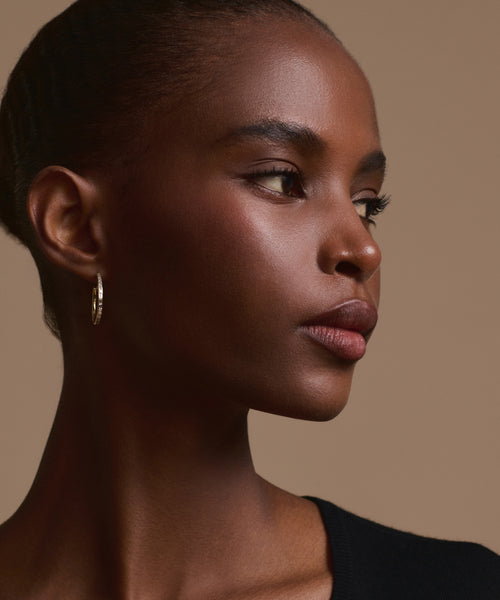 A woman with smooth dark skin and braided hair looks to the side, wearing the June Baguette Hoops in 14k gold and a black top, against a plain light tan background.