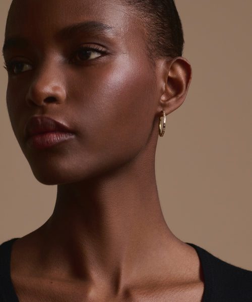 A close-up of a woman with deep brown skin wearing Jenni Kayne June Baguette Hoops. She has short hair, a neutral expression, and is dressed in a black top against a beige background.