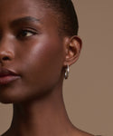 A close-up of a person with smooth dark skin wearing our elegant June Baguette Hoops in 14k gold, set against a plain beige background. Only the left side of their face and neck is visible.