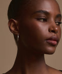 A close-up of a woman with dark skin and short hair, gazing to the side with a calm expression. She wears June Baguette Hoops in 14k gold and natural makeup, set against a neutral background.