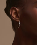Close-up of a person's ear wearing Jenni Kayne's June Baguette Hoops—a small silver hoop earring—against smooth dark skin and pulled-back hair, set against a plain beige background.