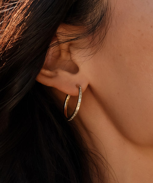 A close-up of a persons ear wearing June Baguette Hoops in 14k gold with a row of sparkling stones. Their dark hair frames the scene, and smooth, evenly lit skin highlights the elegant detail of these diamond earrings.