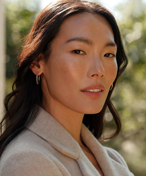 A woman with long dark hair in a beige coat and June Baguette Hoops stands confidently outdoors, sunlight highlighting her against a softly blurred green background.