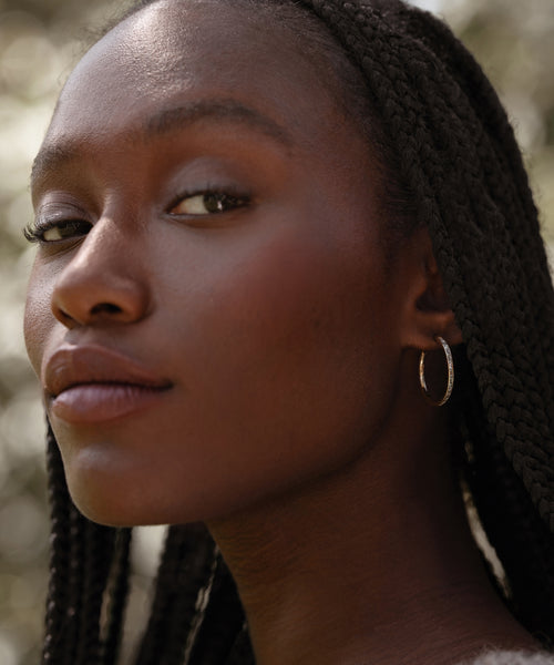 A close-up of a woman with braided hair, wearing June Baguette Hoops, gazes confidently at the camera. The softly blurred background highlights her serene expression and elegant 14k gold June Baguette Hoops.