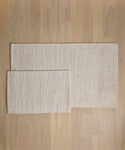 Two rectangular beige Wilder Jute Rugs with a textured, handwoven surface are placed on a light wooden floor, with the smaller rug partially overlapping the larger one from the left.