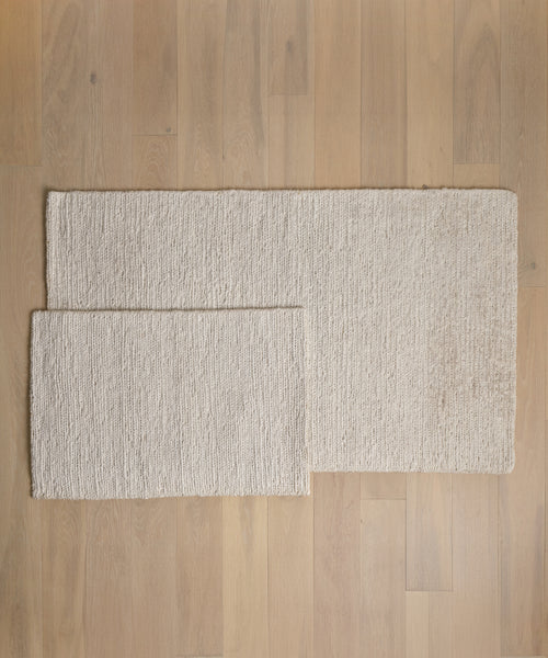 Two rectangular beige Wilder Jute Rugs with a textured, handwoven surface are placed on a light wooden floor, with the smaller rug partially overlapping the larger one from the left.