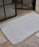 The Wilder Jute Rug, in light gray, is handwoven and lies on a stone tile floor in front of a partially open black-framed glass door.