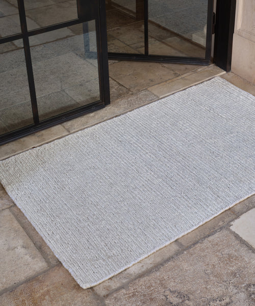 The Wilder Jute Rug, in light gray, is handwoven and lies on a stone tile floor in front of a partially open black-framed glass door.