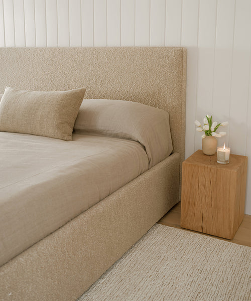 A neatly made bed with beige bedding and headboard sits by a wooden nightstand with a vase of white flowers and a lit candle, set against a white paneled wall and anchored by the Wilder Jute Rug.