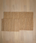 Two Wilder Jute Rugs in different sizes are placed on a light wooden floor, with the smaller rug partially overlapping the larger one. Both rugs showcase a natural, textured look.