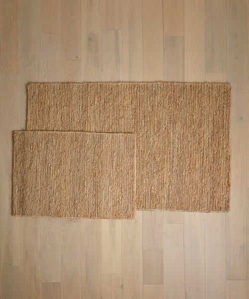 Two Wilder Jute Rugs in different sizes are placed on a light wooden floor, with the smaller rug partially overlapping the larger one. Both rugs showcase a natural, textured look.