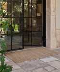 A glass door is partially open, revealing an outdoor stone patio with a large rectangular Wilder Jute Rug. Nearby, green plants and architectural details are visible.