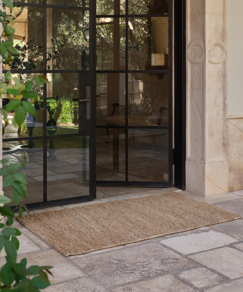 A glass door is partially open, revealing an outdoor stone patio with a large rectangular Wilder Jute Rug. Nearby, green plants and architectural details are visible.