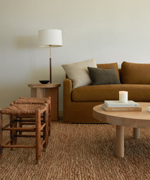 A cozy living room with a brown sofa, neutral pillows, the Wilder Jute Rug, a wooden coffee table with books and a lit candle, a side table with a lamp, and two woven stools. The minimalist decor feels earthy and inviting.