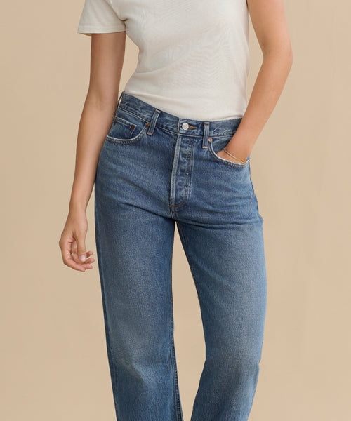 Wearing the Kelly Jean, a person stands with one hand in their pocket, pairing high-rise straight jeans with a white short-sleeve tee for an effortlessly cool, sustainable denim look against a beige background.