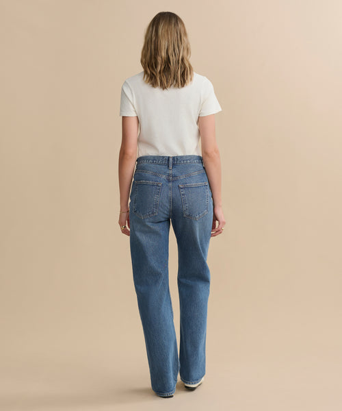 A person with shoulder-length hair faces away, wearing a white short-sleeve tee and the Kelly Jean high-rise straight jeans, crafted from sustainable denim, set against a plain beige background.