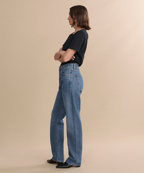 A person with shoulder-length brown hair stands in profile against a beige background, wearing a black t-shirt, the Kelly Jean high-rise straight jeans, and black shoes with arms crossed.