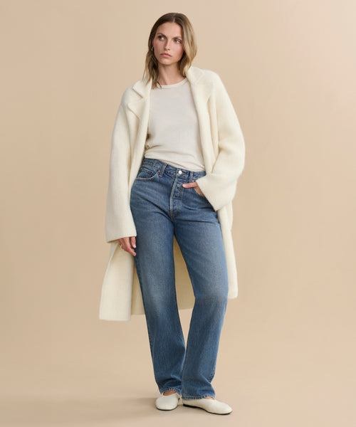 A woman in a long cream coat, light sweater, and white shoes styles the Kelly Jean—high-rise straight jeans—for a timeless look, standing against a beige background with a neutral expression as she gazes to the side.