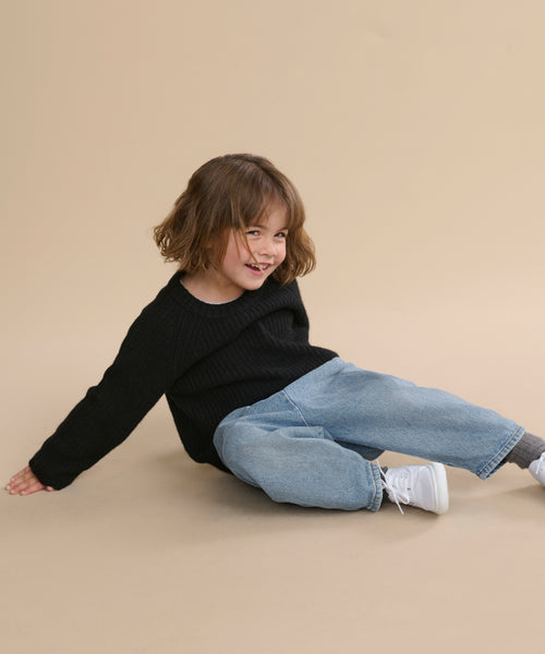 A smiling young child with short brown hair wears a black Kids' Cashmere Fisherman sweater, loose blue jeans, and white sneakers while sitting on a beige floor against a matching beige background.