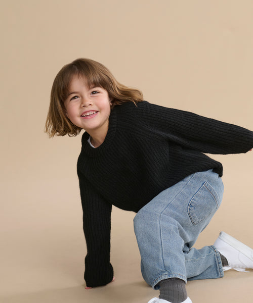 A young child with shoulder-length brown hair, in a black Kids' Cashmere Fisherman sweater, blue jeans, and white sneakers, crouches and smiles against a plain beige background.