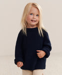 A young child with long blonde hair and blue eyes smiles while wearing the Kids Cashmere Fisherman sweater in navy and light-colored pants, standing against a neutral background.