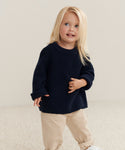 A young child with long blonde hair stands on a light carpet, smiling and glancing to the side while wearing beige pants and the Kids Cashmere Fisherman sweater in navy.