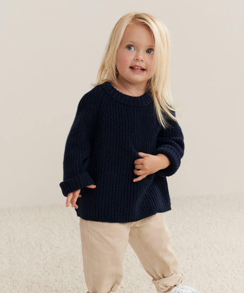A young child with long blonde hair stands on a light carpet, smiling and glancing to the side while wearing beige pants and the Kids Cashmere Fisherman sweater in navy.