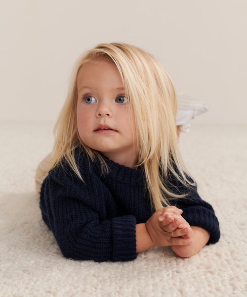 A young child with long blonde hair lies on a soft, light-colored carpet, wearing the cozy Kids Cashmere Fisherman sweater in navy and gazing upward with a thoughtful look.