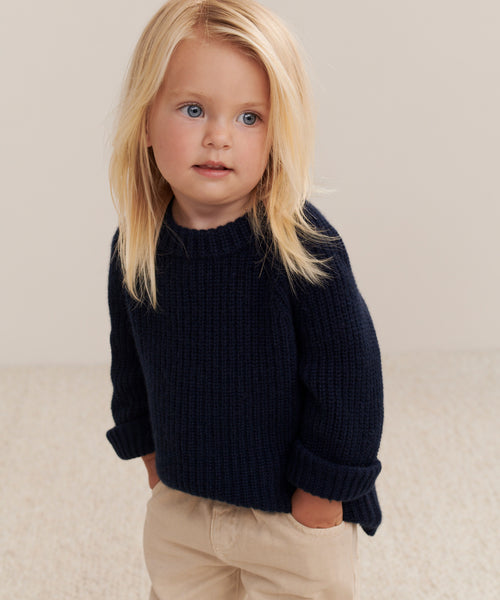 A young child with long blonde hair stands with hands in pockets, wearing the Kids Cashmere Fisherman sweater in navy and beige pants, gazing slightly to the side against a neutral background.