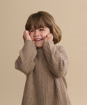 A young child with brown hair smiles joyfully, hands on their cheeks, while wearing a cozy Kids' Cashmere Fisherman sweater against a plain beige background.