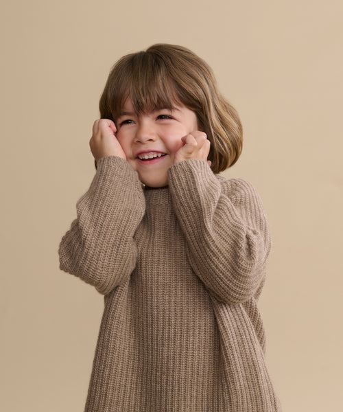 A young child with brown hair smiles joyfully, hands on their cheeks, while wearing a cozy Kids' Cashmere Fisherman sweater against a plain beige background.