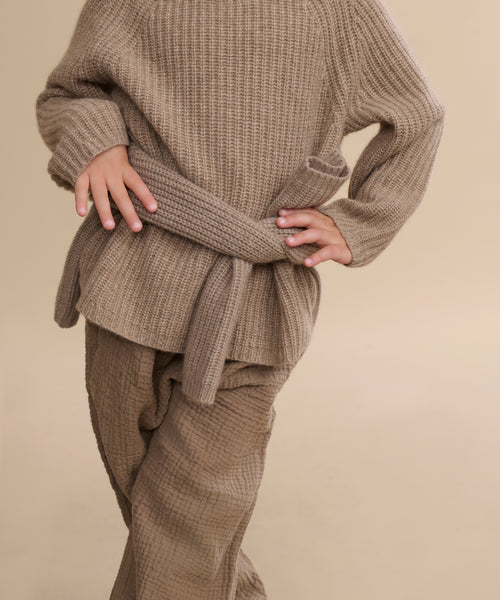 A child wears the Kids' Cashmere Fisherman sweater in undyed stone, styled oversized with rolled-up sleeves and matching wide-leg pants, standing hands on hips against a plain beige background.