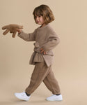 A young child with wavy brown hair walks against a beige background, wearing the Kids' Cashmere Fisherman sweater and white sneakers, holding a brown stuffed animal.