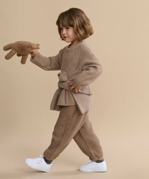 A young child with wavy brown hair walks against a beige background, wearing the Kids' Cashmere Fisherman sweater and white sneakers, holding a brown stuffed animal.