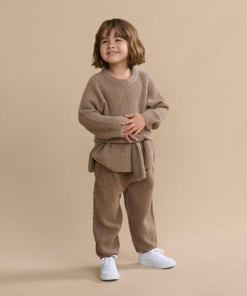 A young child with short brown hair smiles, wearing the cozy undyed stone Kids' Cashmere Fisherman sweater and matching pants with white sneakers, standing against a plain beige background.