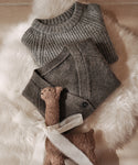 Two folded heather grey knit sweaters, one being the Kids Cashmere Fisherman, rest on a fluffy white surface with a small brown plush toy llama tied with a white ribbon in front.