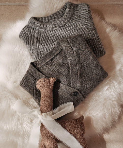 Two folded heather grey knit sweaters, one being the Kids Cashmere Fisherman, rest on a fluffy white surface with a small brown plush toy llama tied with a white ribbon in front.
