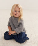 A young child with long blonde hair, wearing a heather grey Kids Cashmere Fisherman sweater and blue jeans, sits on a textured cream-colored carpet, smiling softly and gazing slightly to the side.