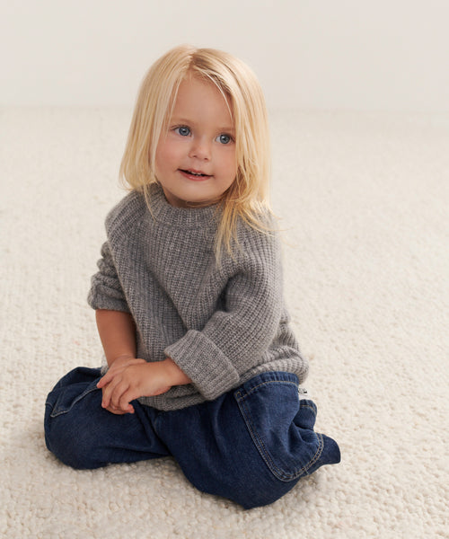 A young child with long blonde hair, wearing a heather grey Kids Cashmere Fisherman sweater and blue jeans, sits on a textured cream-colored carpet, smiling softly and gazing slightly to the side.