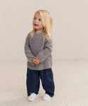 A young child with long blonde hair wears the Kids Cashmere Fisherman sweater in heather grey, paired with blue jeans and white shoes, standing on a light carpet against a beige background, smiling and looking up.