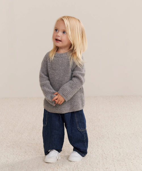 A young child with long blonde hair wears the Kids Cashmere Fisherman sweater in heather grey, paired with blue jeans and white shoes, standing on a light carpet against a beige background, smiling and looking up.